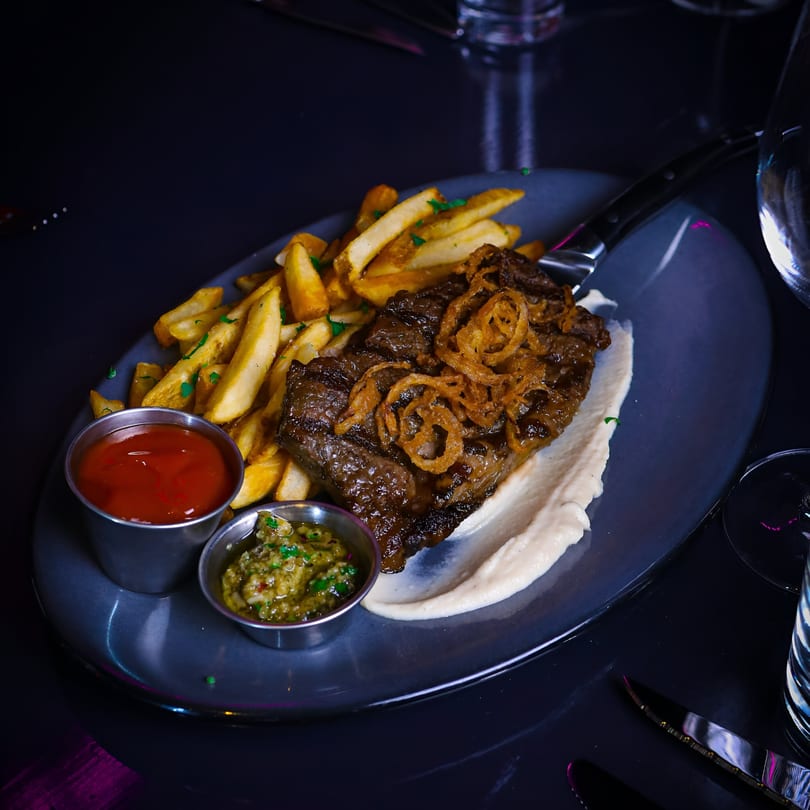 New-American steak frites with crispy onions, a white sauce, ketchup, and relish on a dark plate.