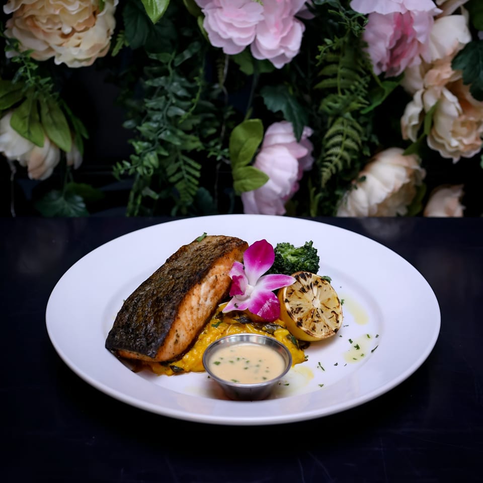 Seared salmon with grilled lemon, broccoli, sauce, and a pink orchid on a white plate.