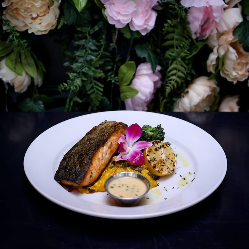 Seared salmon with grilled lemon, broccoli, sauce, and a pink orchid on a white plate.