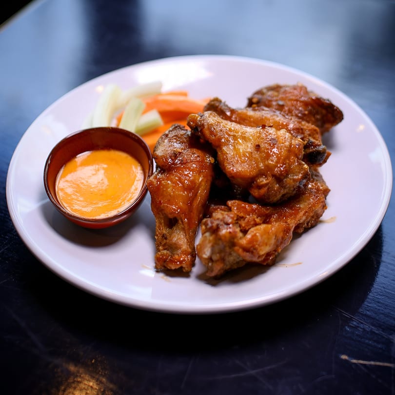 Glazed chicken wings with a side of dipping sauce, celery, and carrot sticks on a white plate.