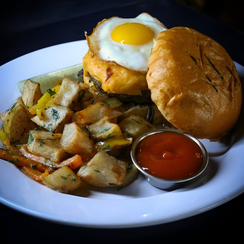 New-American burger with fried egg, cheese, and a side of roasted potatoes and ketchup.