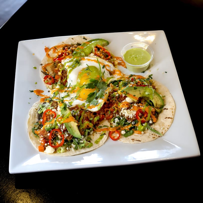 Three breakfast tacos with fried eggs, avocado, red peppers, and green sauce on a white plate.