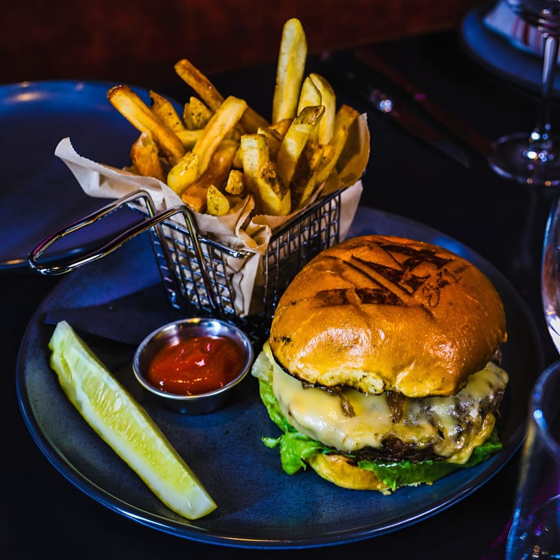 A V 14 Street Lounge cheeseburger with fries, ketchup, and a pickle spear on a dark plate.