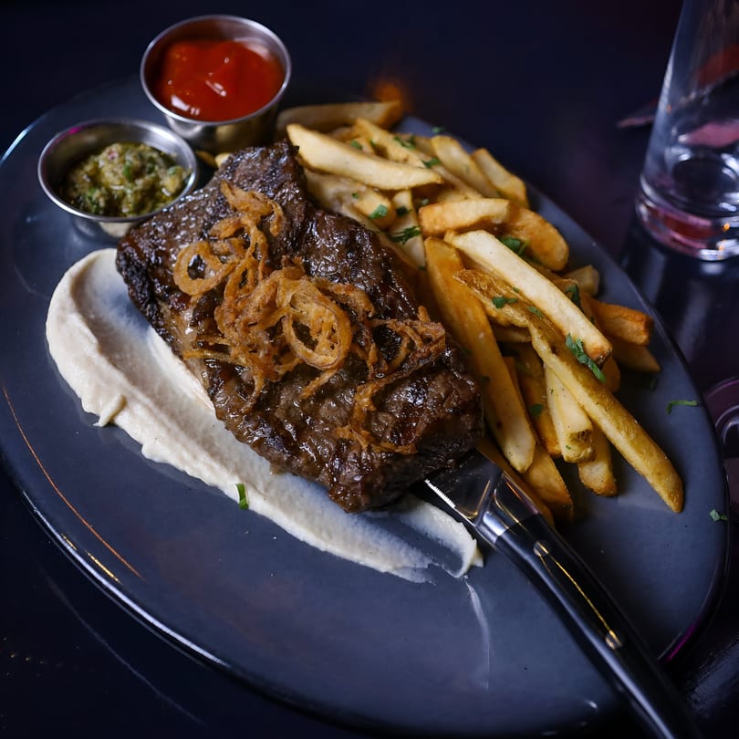 New-American steak with crispy onions, fries, and two dipping sauces on a dark plate.